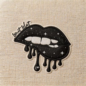 May include: Embroidered patch featuring black lips with white teeth and dripping details. The lips are adorned with stars and the text "smut slut" in cursive. The patch is on a beige fabric background.