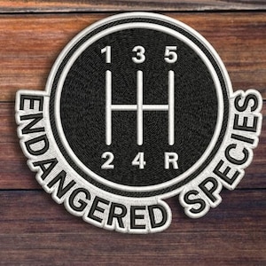 May include: Embroidered patch featuring a black gear shift pattern with white numbers and lines, encircled by a white border. The words "ENDANGERED SPECIES" are embroidered around the circle in white. The patch is set against a wooden background.