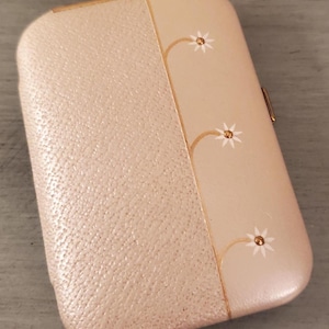 May include: A vintage, rectangular, blush-colored case with a textured finish on one side and a smooth, cream-colored panel on the other. The cream panel features a gold-lined floral design with white star-shaped accents. The case has a gold clasp.