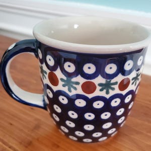 May include: A ceramic mug featuring a dark blue background with a repeating pattern of white dots, dark blue circles, reddish-brown circles, and small green star-like shapes. The mug has a curved handle and a smooth, cream-colored interior rim.