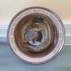 May include: A round ceramic bowl with a wide, cream-colored rim featuring the black text, "MAY YOUR HOME BE FILLED WITH HAPPINESS, TRANQUILITY AND FRIENDSHIP." The interior of the bowl displays a swirling, multi-colored glaze in shades of brown, blue, and red.