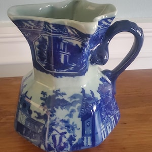 May include: A blue and white ceramic pitcher with a detailed landscape design. The pitcher has a unique shape with a flared top and a curved handle. The design features buildings, trees, and other elements in a classic blue and white color scheme.