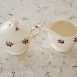 May include: Cream-colored ceramic sugar bowl with lid and creamer set. Both pieces feature floral accents in red, blue, and purple. The sugar bowl has a lid with a flower-shaped handle. The creamer has a spout and handle.