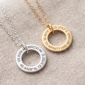 May include: Two circular pendant necklaces on delicate chains. One pendant is silver-toned with engraved text reading VII.XVIII.MXCVI, an infinity symbol, and 45.4408 N, 12.3155 E. The other is gold-toned with engraved text reading 25.3753 E and TO INFINITY AND BEYOND.