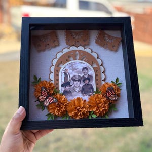 May include: A black shadow box frame containing a central oval photograph of five individuals, surrounded by layered paper marigold flowers, two monarch butterflies, and three decorative paper banners against a light gray fabric background.