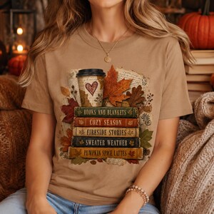 Woman wearing a light brown fall book lover shirt featuring stacked vintage books, coffee cup, autumn leaves, cozy season and sweater weather design.