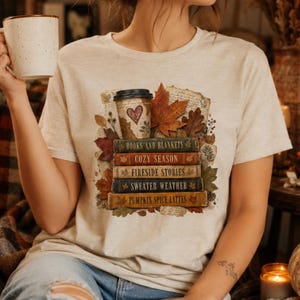 Woman wearing a cream fall book lover shirt featuring autumn leaves, stacked books, coffee cup, cozy season and pumpkin spice latte design.