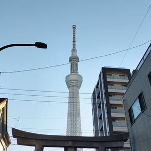 以下が含まれることがあります： 東京スカイツリーの低いアングルからの写真。高い電波塔で、複雑な格子構造が特徴。青い空を背景に、周囲の建物や電線が風景を構成しています。