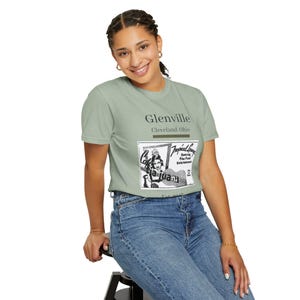 May include: A light green t-shirt with the word "Glenville" and "Cleveland, Ohio" printed on the front. Below is a black and white graphic of a woman and the words "Cafe La Juana" and "Tropical Lounge".