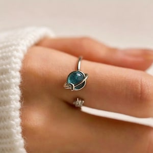 Könnte beinhalten: Ein silberfarbener offener Ring mit einem kleinen, runden, durchscheinenden blauen Stein in der Mitte, der von einem dünnen Metallring umgeben ist, der an einen Planeten erinnert, sowie einem winzigen sternförmigen Anhänger am Ende.