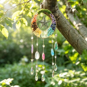 May include: A circular metal suncatcher featuring a crescent moon shape filled with multicoloured gemstone chips in a rainbow gradient. A silver star hangs in the centre, topped with green moss. Five dangling chains feature crystal teardrops.