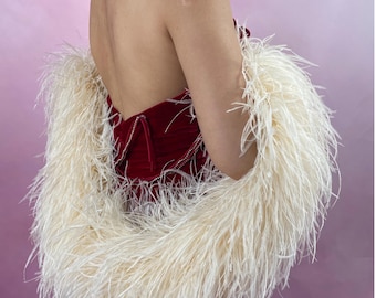 May include: A champagne-15ply ostrich feather boa draped around a deep red velvet strapless garment. The boa consists of long, thin, off-white feathers that create a voluminous, textured appearance against the smooth, dark red fabric of the dress.