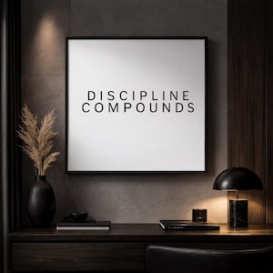 Könnte beinhalten: Ein gerahmter Druck mit den Worten "DISCIPLINE COMPOUNDS" in schwarzer Schrift. Der Druck hängt über einem dunklen Holzschreibtisch mit einer schwarzen Lampe, Vase und anderen Dekorationen. Die Gesamtästhetik ist modern und minimalistisch.