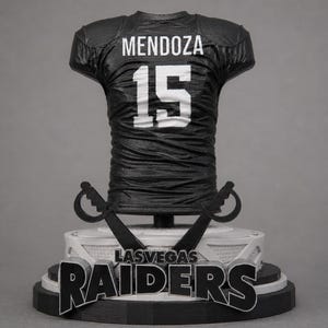 Op de afbeelding: Een zwart-witte sculptuur van een Las Vegas Raiders voetbalshirt. Het shirt toont de naam "MENDOZA" en het nummer "15" in het wit. De basis toont de teamnaam en het logo, met gekruiste zwaarden.