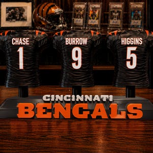May include: A black Cincinnati Bengals display featuring three miniature football jerseys with the names Chase, Burrow, and Higgins. The jerseys have white numbers and lettering. The base reads "CINCINNATI BENGALS" in white and orange.
