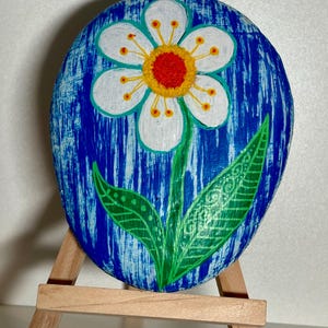 May include: An oval painted stone with a white flower, red centre, and yellow details, set against a blue backdrop. The flower has green leaves and stem. The stone is displayed on a small wooden easel.