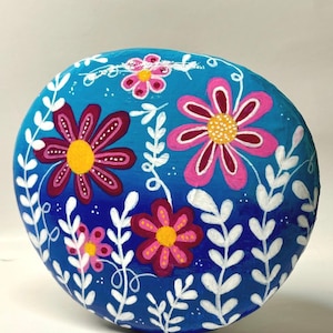 May include: A round, smooth stone painted with a vibrant blue background featuring several hand-painted flowers in shades of pink, magenta, and yellow. The design includes delicate white leafy vines and small white dots scattered across the surface.