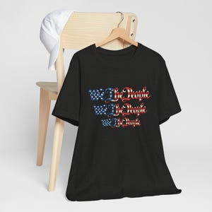 Könnte beinhalten: Schwarzes T-Shirt mit dem Text "We The People" in Rot und Blau, mit einem Design, das an die amerikanische Flagge erinnert. Das Shirt hängt an einem Holzkleiderbügel. Eine weiße Baseballkappe liegt auf einem Holzstuhl.
