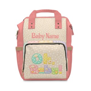 以下が含まれることがあります： ピンクとベージュのおむつバッグ。テキスト「Baby Name」と「Oh, Baby!」が遊び心のあるフォントで書かれています。バッグは水玉模様で、カラフルな刺繍文字と画像が特徴です。上部にハンドルとサイドポケットが付いています。