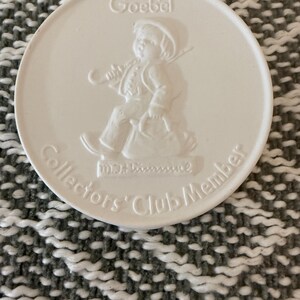 May include: A white ceramic Goebel Collectors Club Member medallion. The medallion features a bas-relief image of a child with a walking stick and a bag. The Goebel logo and text are embossed on the surface.