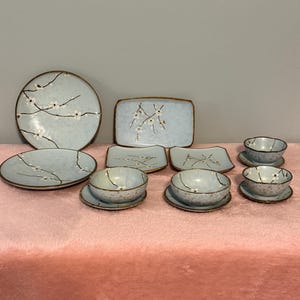 May include: A collection of light blue ceramic tableware with a floral design. The set includes plates, bowls, and saucers. The edges of the plates and bowls have a brown trim. The floral design features white blossoms and thin black branches.