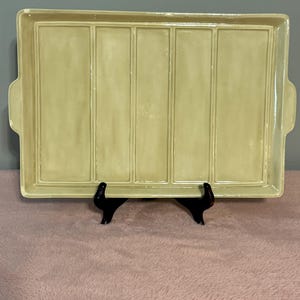 May include: A rectangular, pale yellow ceramic serving tray with a divided design. The tray has a raised rim and two small handles. The tray is supported by a black stand, and is displayed on a pink surface.