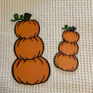 May include: Two stacked pumpkin decorations made of orange material with dark brown outlines and small green vine accents at the top. The larger pumpkin stack consists of three rounded segments, while the smaller stack also features three segments.
