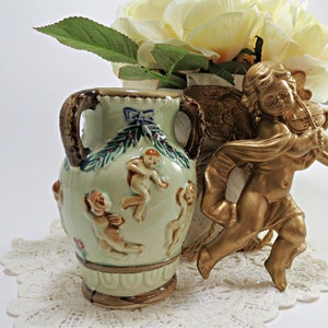 May include: A small, green ceramic vase with two handles and a floral design. The vase is decorated with cherubs and a bow. It is sitting on a white lace doily next to a gold cherub figurine playing a violin.