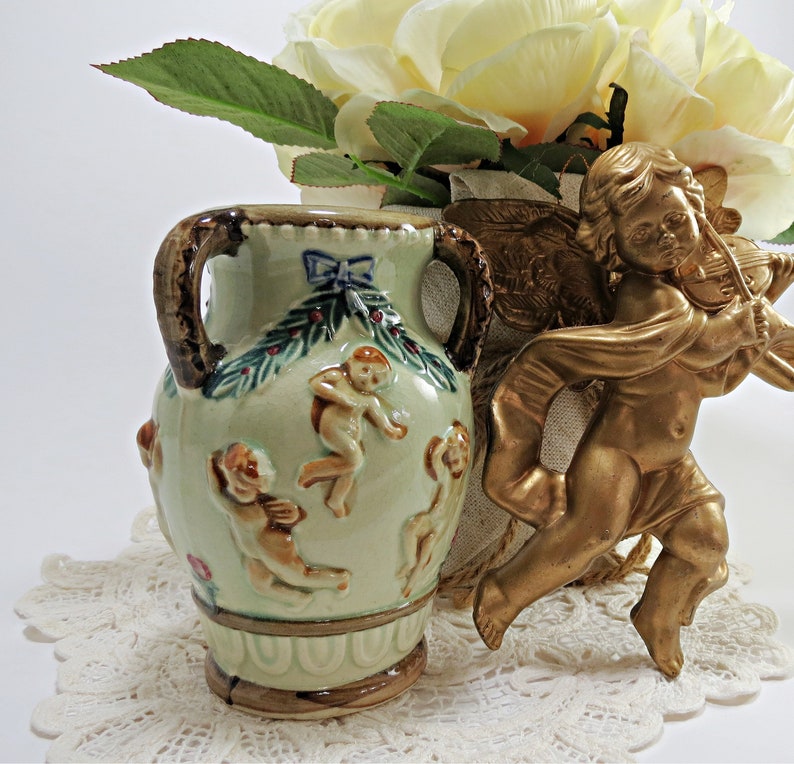 May include: A small, green ceramic vase with two handles and a floral design. The vase is decorated with cherubs and a bow. It is sitting on a white lace doily next to a gold cherub figurine playing a violin.