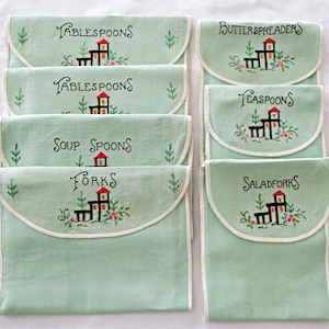 May include: A set of vintage embroidered utensil holders in a soft green hue. Each holder is labeled with utensil names like "Tablespoons" and "Forks", and features a charming house design with floral accents. The holders have a white trim.