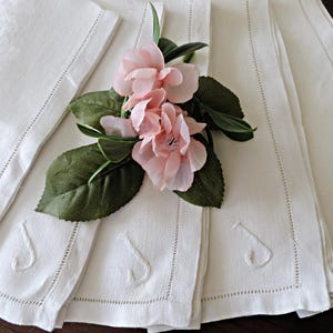 May include: White linen napkins with embroidered details, arranged with pink flowers and green leaves. The napkins have a decorative stitched border and an embroidered design. The image showcases a set of elegant table linens.