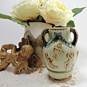 May include: A small, green ceramic vase with two handles and a design of cherubs and greenery. The vase is decorated with brown and gold accents.
