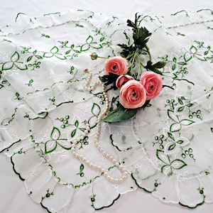 May include: White embroidered table runner with green floral detailing and scalloped edges. The runner is adorned with pink roses and a pearl necklace, creating an elegant and decorative display.