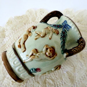 May include: A small, green ceramic vase with brown handles and a relief design of cherubs. The vase has a blue bow and green leaves around the top.