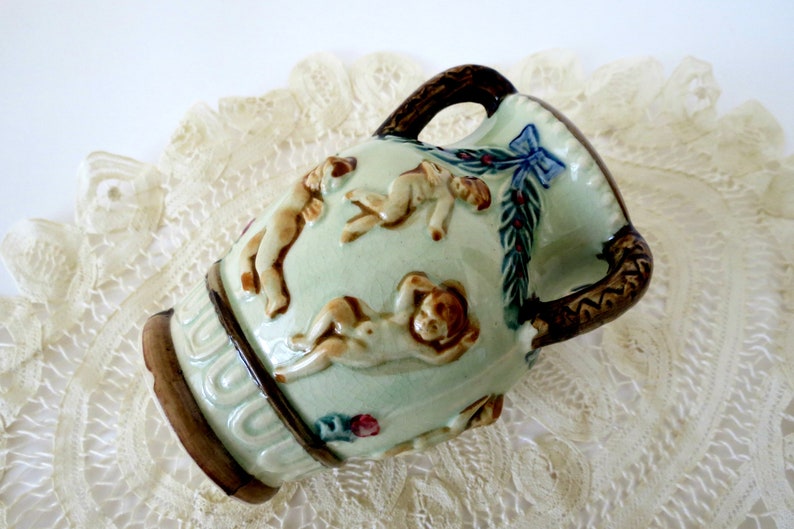 May include: A small, green ceramic vase with brown handles and a relief design of cherubs. The vase has a blue bow and green leaves around the top.