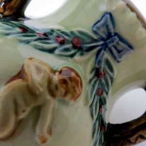 May include: A ceramic pitcher with a green and brown glaze. The pitcher features a sculpted design of a cherub, a bow, and a garland of leaves and berries.