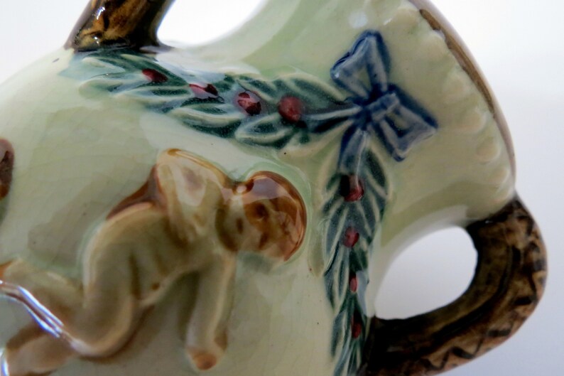 May include: A ceramic pitcher with a green and brown glaze. The pitcher features a sculpted design of a cherub, a bow, and a garland of leaves and berries.