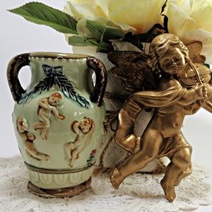 May include: A green ceramic vase with two handles and a design of cherubs playing music. The vase is sitting on a white lace doily next to a gold cherub figurine playing a violin.
