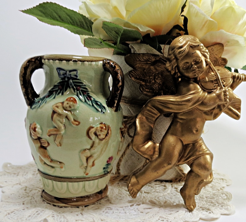 May include: A green ceramic vase with two handles and a design of cherubs playing music. The vase is sitting on a white lace doily next to a gold cherub figurine playing a violin.