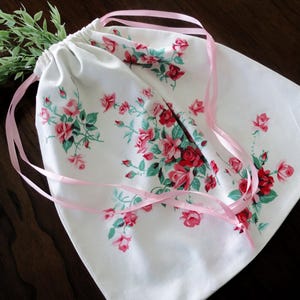 May include: White drawstring bag with a floral rose pattern in red and pink, accented with green leaves. The bag has pink ribbon drawstrings. A sprig of green leaves is placed near the top of the bag.