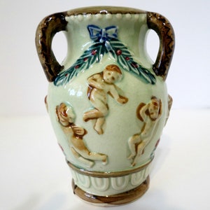 May include: A small, green ceramic vase with two brown handles. The vase is decorated with cherubs, a green wreath, and red berries.