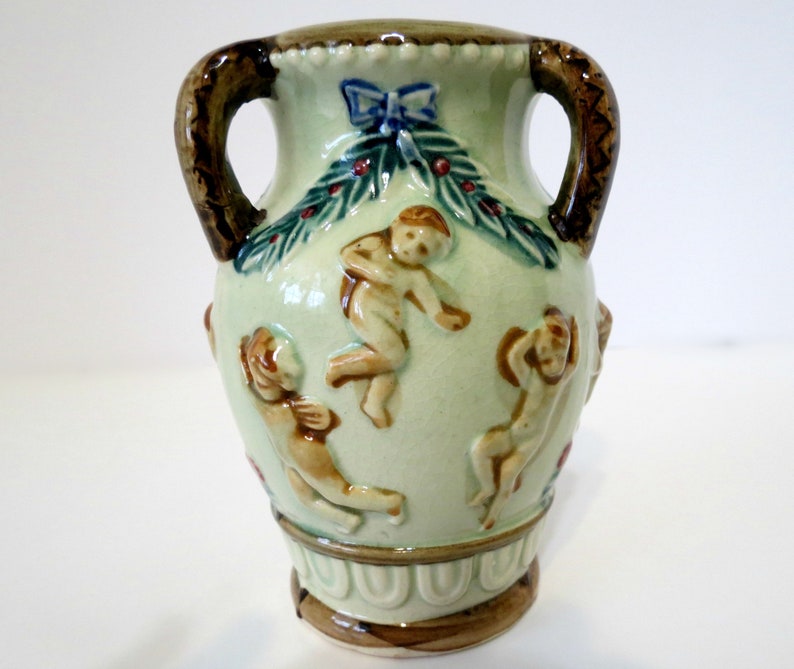 May include: A small, green ceramic vase with two brown handles. The vase is decorated with cherubs, a green wreath, and red berries.