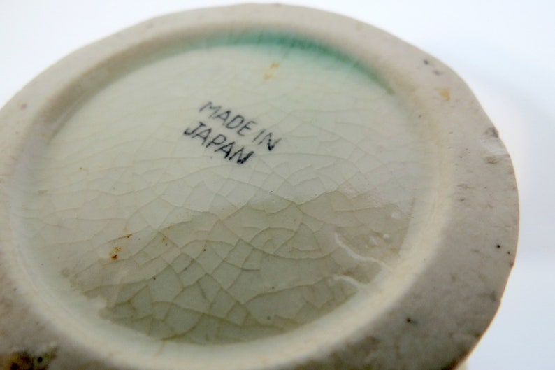 May include: Close-up of the bottom of a white ceramic object with a green glaze. The bottom has a crackled glaze and the words "Made in Japan" are printed in black.
