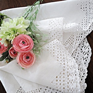May include: A white fabric napkin featuring an intricate crocheted lace border along the edges. A small bouquet of artificial flowers, including pale pink ranunculus and light green foliage, rests on top of the folded white textile.