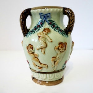 May include: A small, green ceramic vase with two brown handles. The vase is decorated with cherubs, a blue bow, and green leaves.