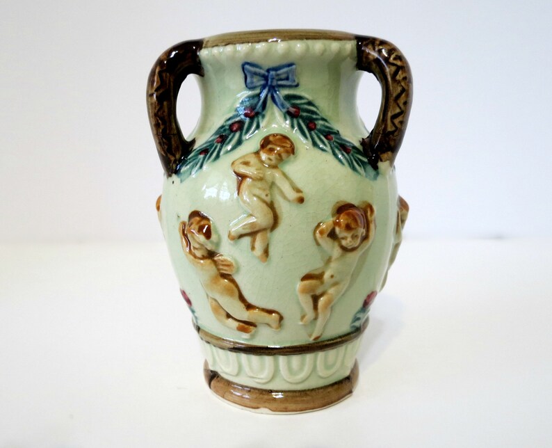 May include: A small, green ceramic vase with two brown handles. The vase is decorated with cherubs, a blue bow, and green leaves.