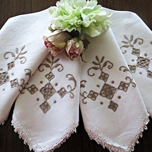 May include: Four white linen napkins with embroidered brown geometric designs and lace edges are arranged on a dark wooden surface. A small bouquet of light green and pink flowers rests on top of the napkins.