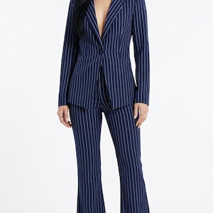 Puede incluir: Traje de raya diplomática azul marino. El traje incluye una blazer con cierre de un solo botón y pantalones acampanados. Las rayas blancas crean un patrón vertical. La modelo lleva tacones negros.