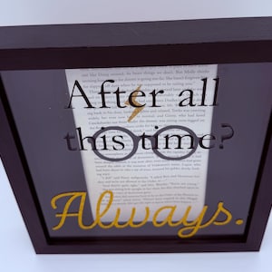 Puede incluir: Una obra de arte enmarcada con un marco de madera marrón oscuro. La obra presenta una página de libro con las palabras "After all this time? Always." en letras negras y doradas, y unas gafas.
