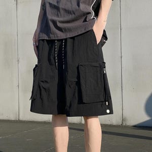 May include: Black knee-length shorts featuring a relaxed fit, an elastic waistband with adjustable drawstring cords, and two large rectangular cargo pockets on the sides. The fabric has a slightly crinkled texture and includes a silver zipper detail.